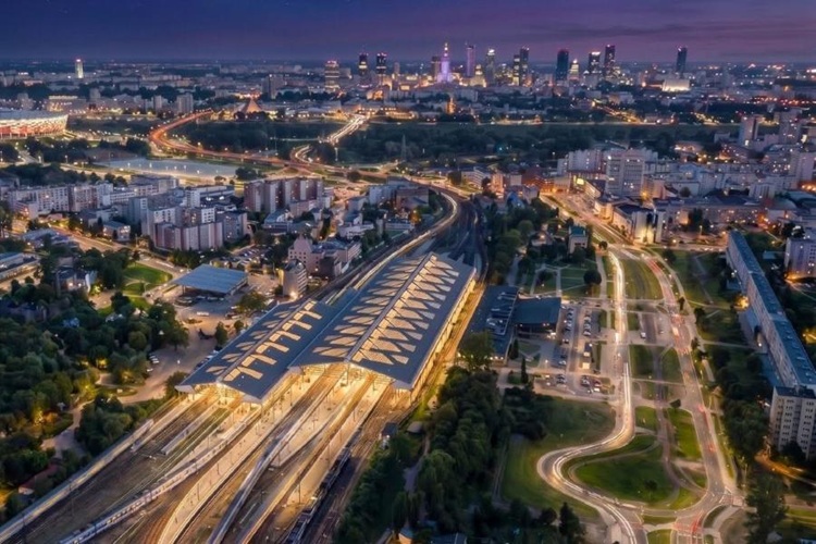 Nocna panorama stacji Warszawa Wschodnia z lotu ptaka — oświetlone tory i perony na tle panoramy Warszawy z Pałacem Kultury i wieżowcami